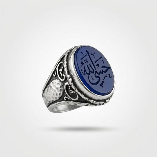 Blauer Jemen Aqīq Ring «Hasbi Allah» aus massivem, gehämmertem 925 Feinsilber mit floralen Schwüngen und einem ovalen, tiefblauen Achat-Cabochon mit der handgravierten arabischen Bittschrift 'حسby الله' (Hasbi Allah), schwebend auf hellem Studio-Hintergrund.